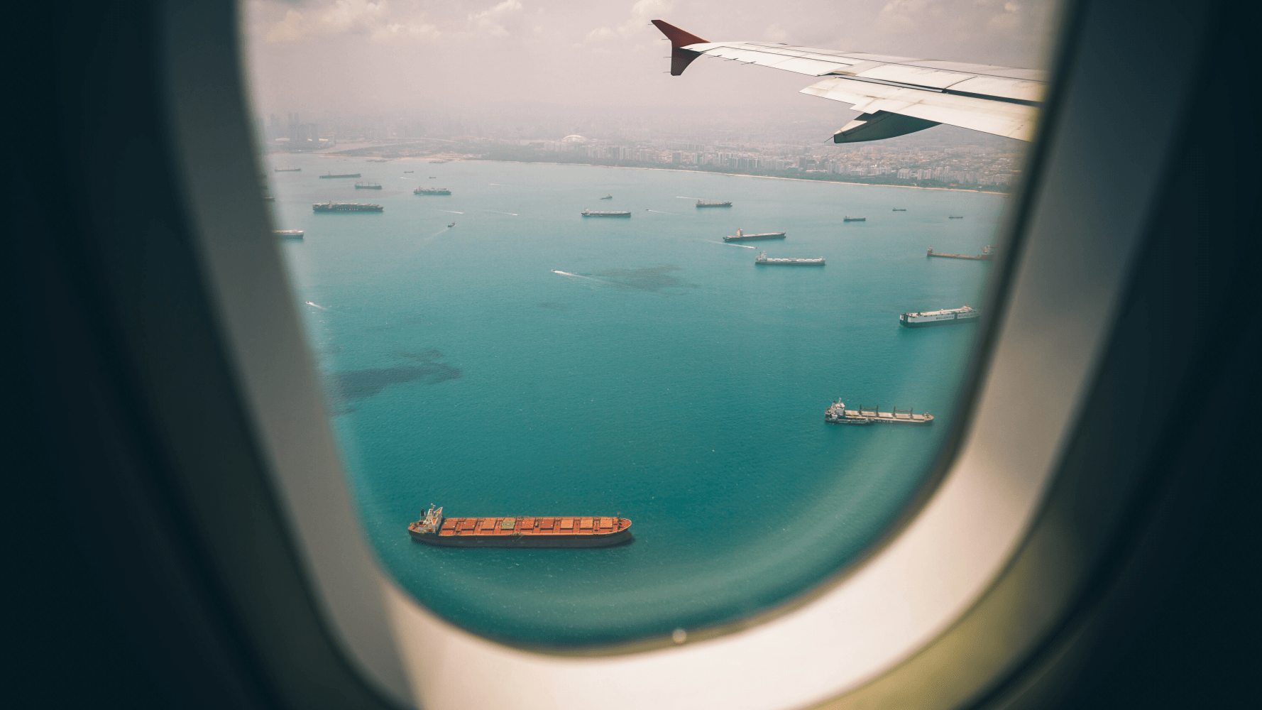 View from the passenger window of a commercial jet overlooking a busy harbor and large container ships